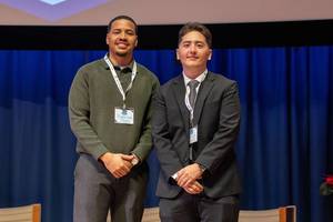Two young adult men in professional attire wearing lanyards with name tags standing on a stage in front of a projector screen 